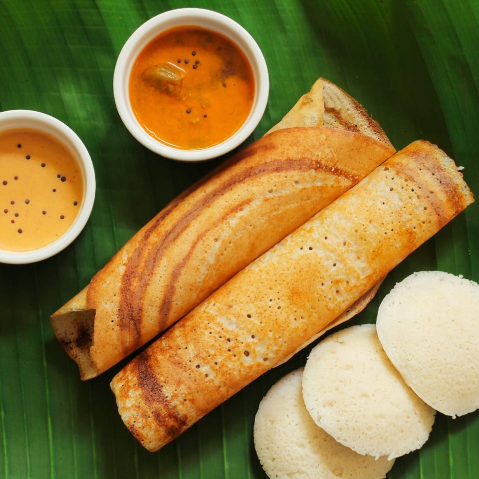 Two golden-brown dosas, two bowls of chutney, and three idlis on a green banana leaf.