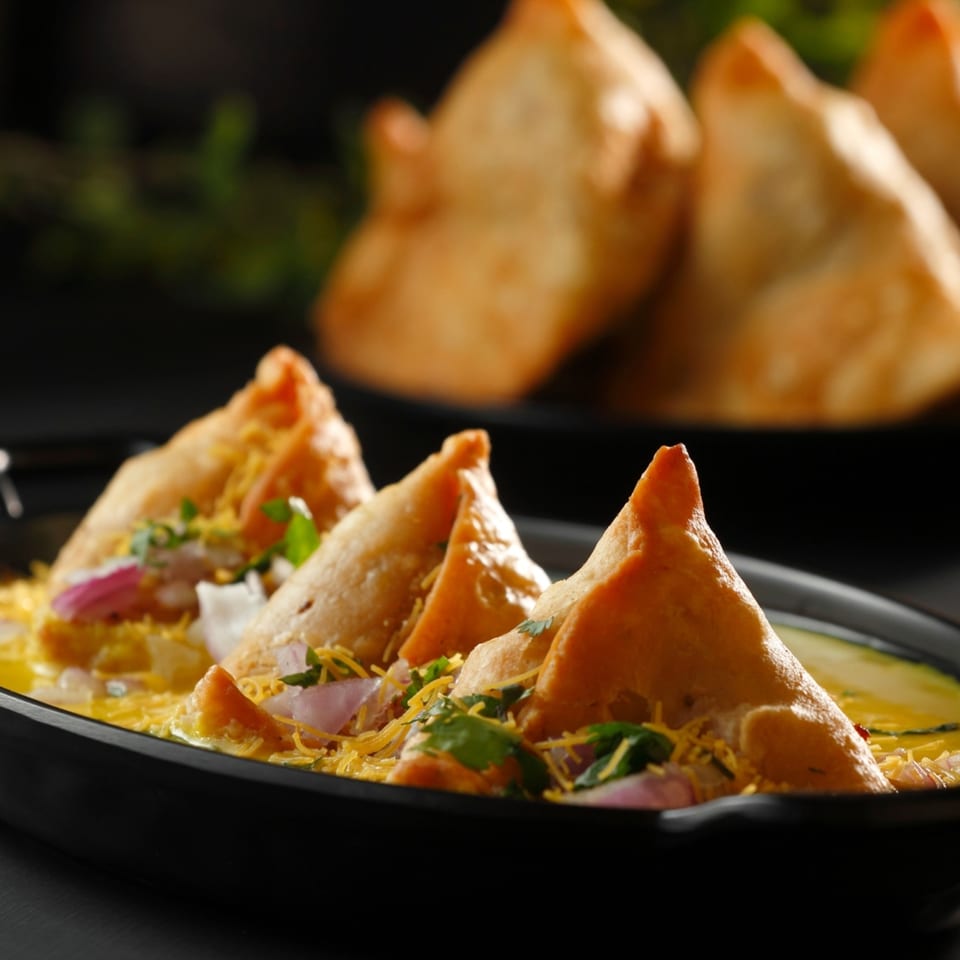 Aha South Indian Cuisine's samosa chaat with crispy samosas, onions, cilantro, and sev in a yellow sauce.