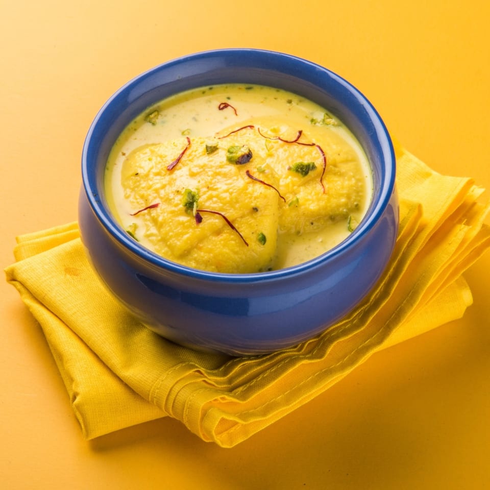 Rasmalai, an Indian dessert, in a blue bowl with saffron and pistachios on a yellow napkin.